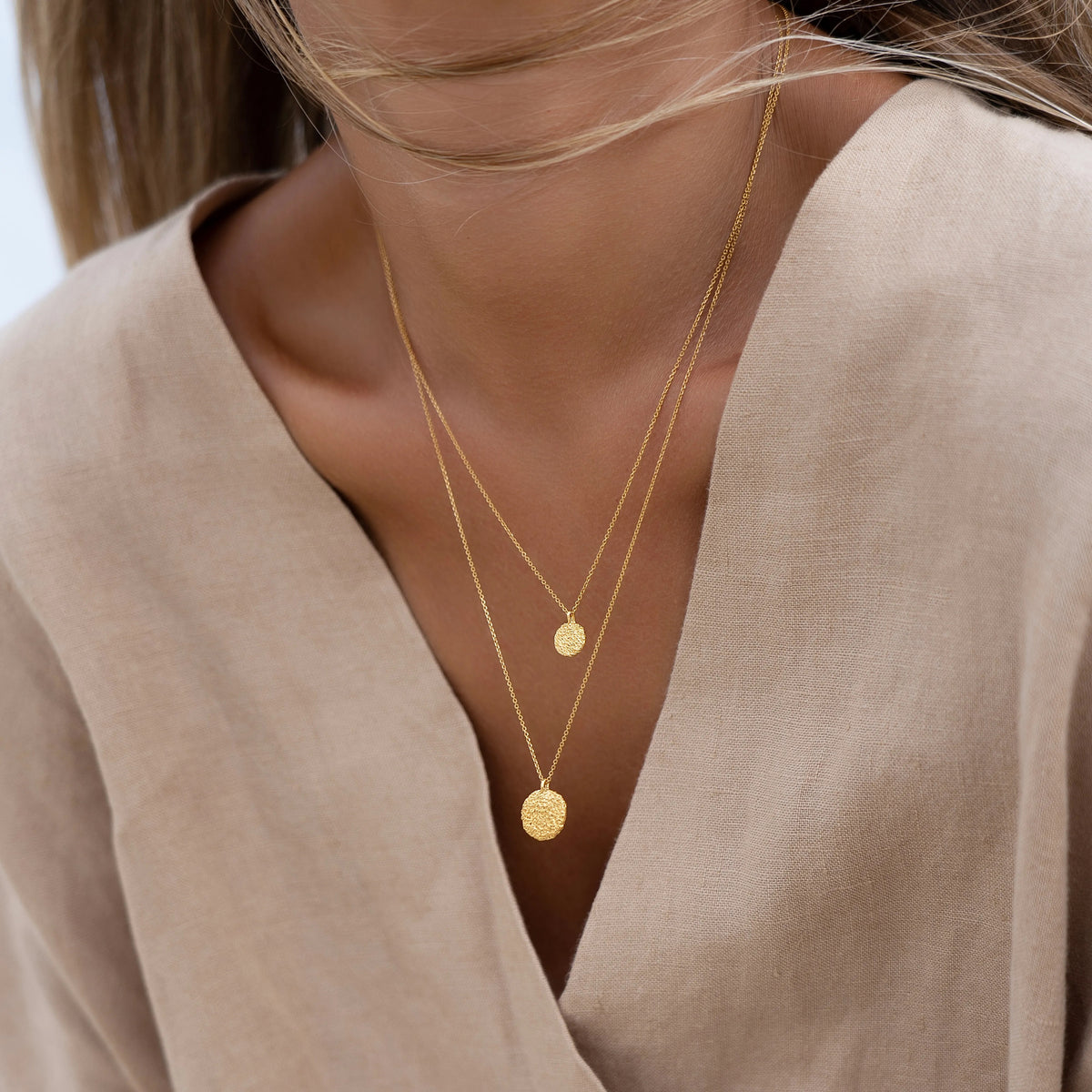 A close-up of a model wearing a double-layered gold necklace with one small flat disc pendant and one textured disc pendant, paired with a beige linen top.