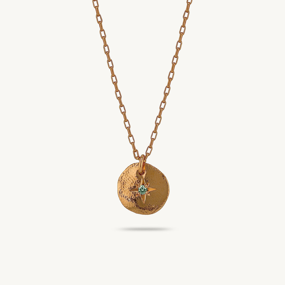 A gold-toned chain necklace with a textured circular pendant featuring a small green starburst detail, shown against a light cream background.