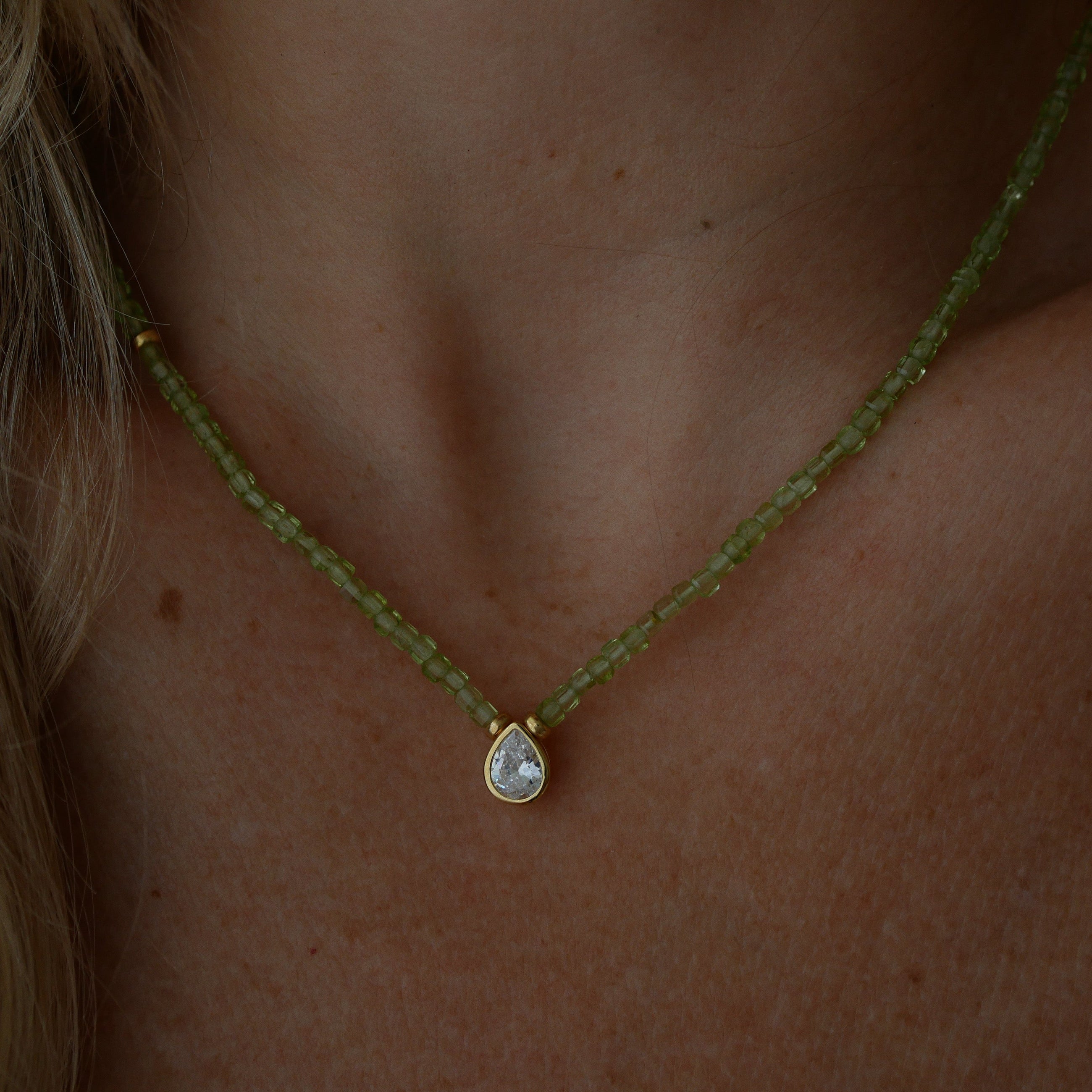 A close-up of a person's neck and chest wearing a delicate necklace featuring a strand of small, faceted peridot beads with a gold-tone setting holding a teardrop-shaped zirconium.
