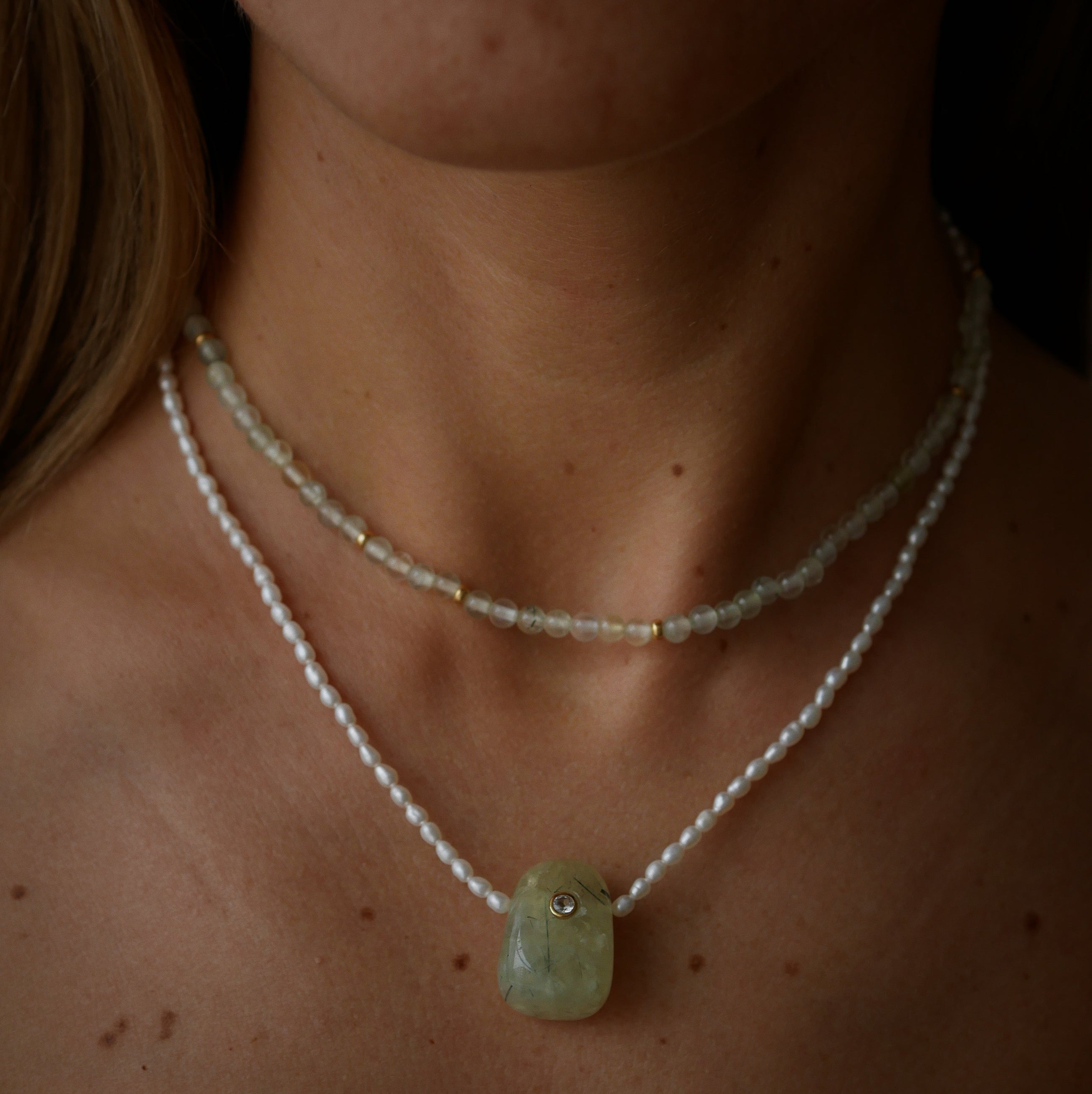A close-up of a person's neck and upper chest wearing two necklaces. Pearl necklace with a light green rectangular pendant and a delicate prehnite beaded strand layered above it.