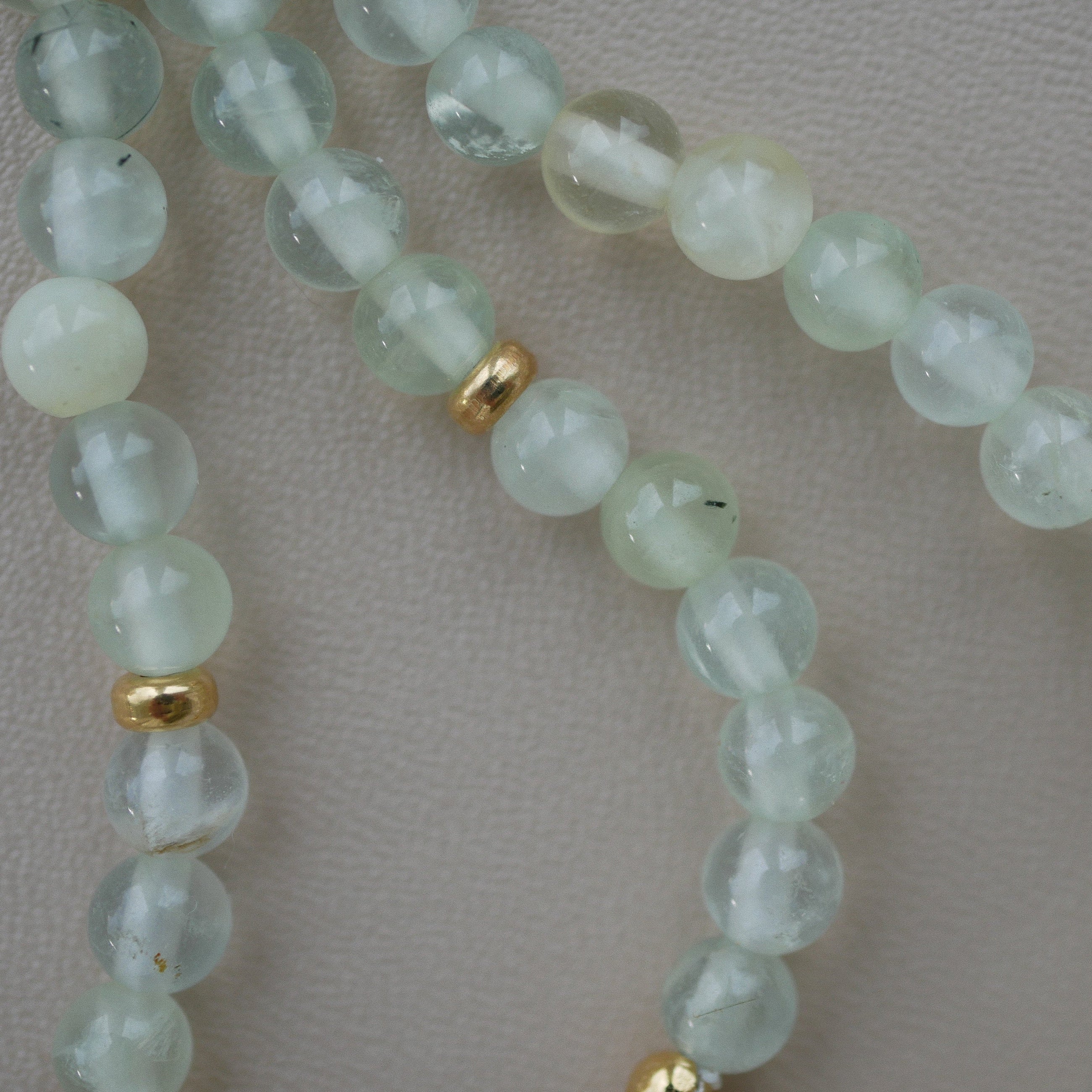 A close-up of a beaded strand featuring pale greenish-white prehnite beads separated by small gold-plated silver spacers, laid on a neutral light beige textured surface.