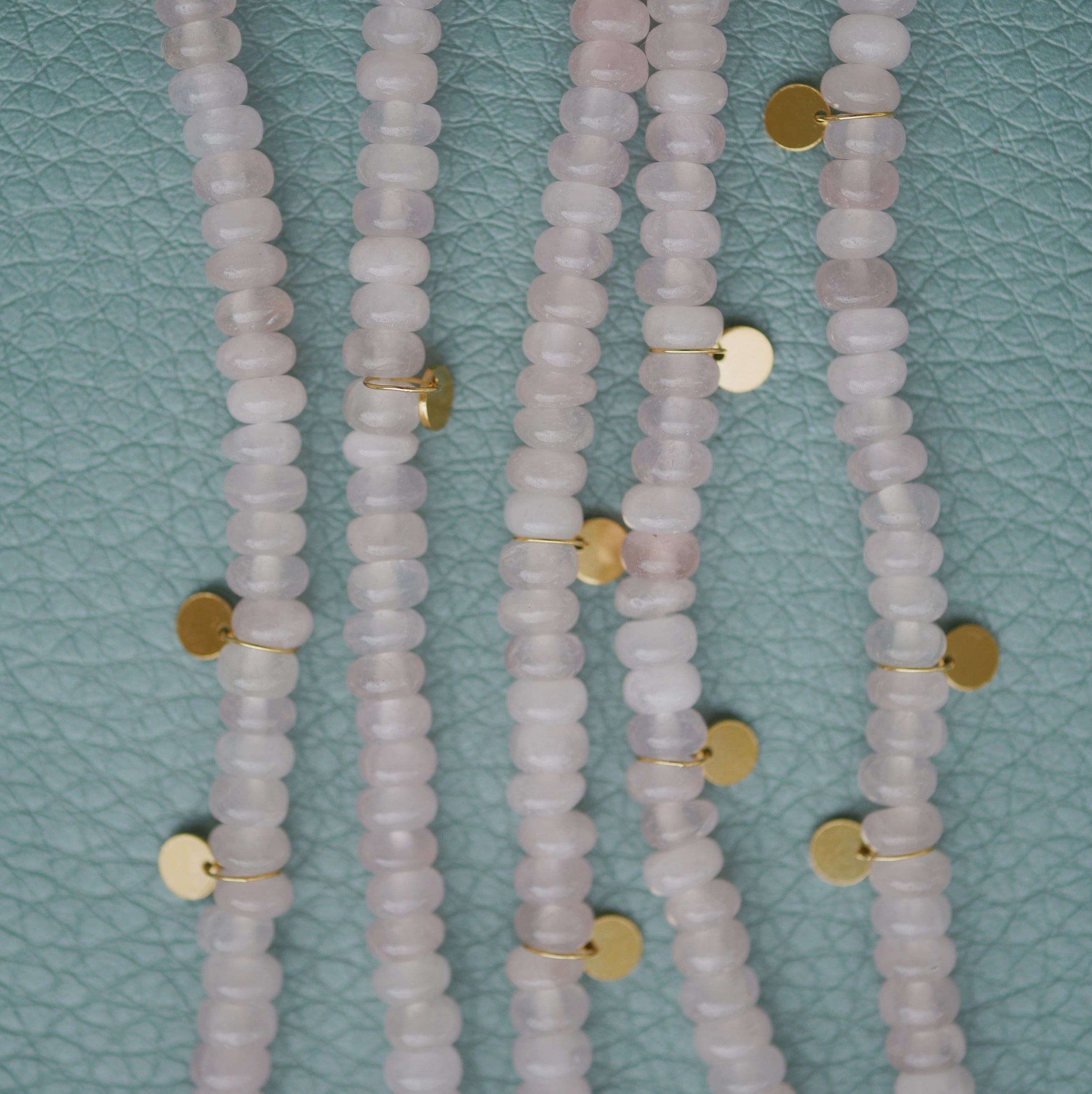 Multiple strands of pale pink quartz beads with small round gold disc charms, laid flat on a textured light blue surface.