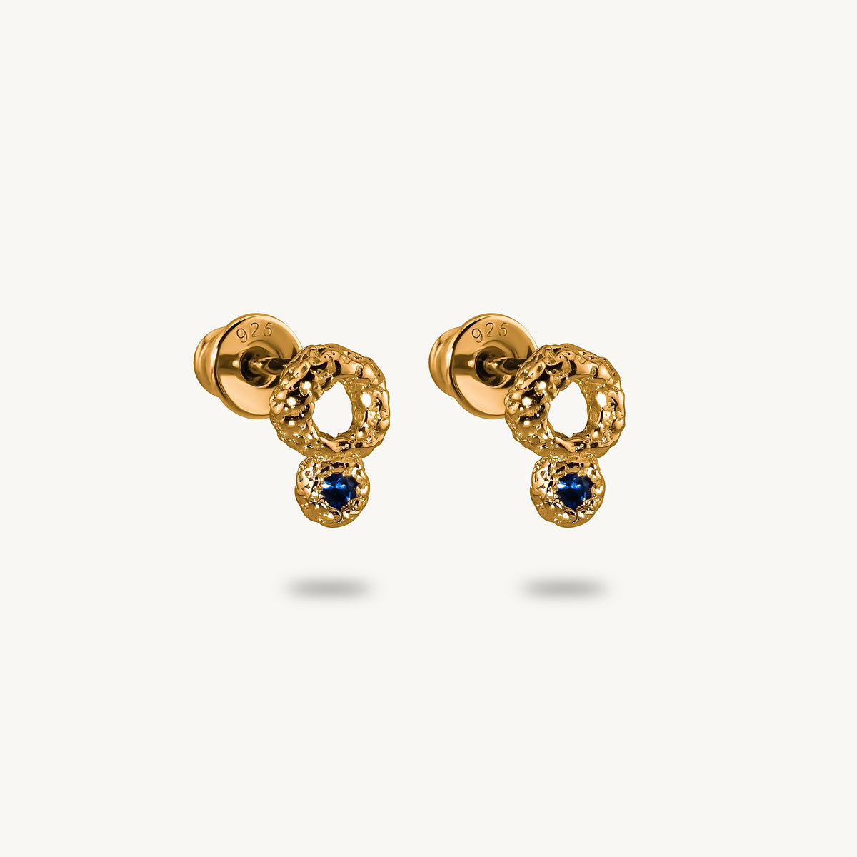 A pair of gold dangle earrings with textured circular loops, small clear pavé accents, and a central round blue sapphire, shown on a light cream background.