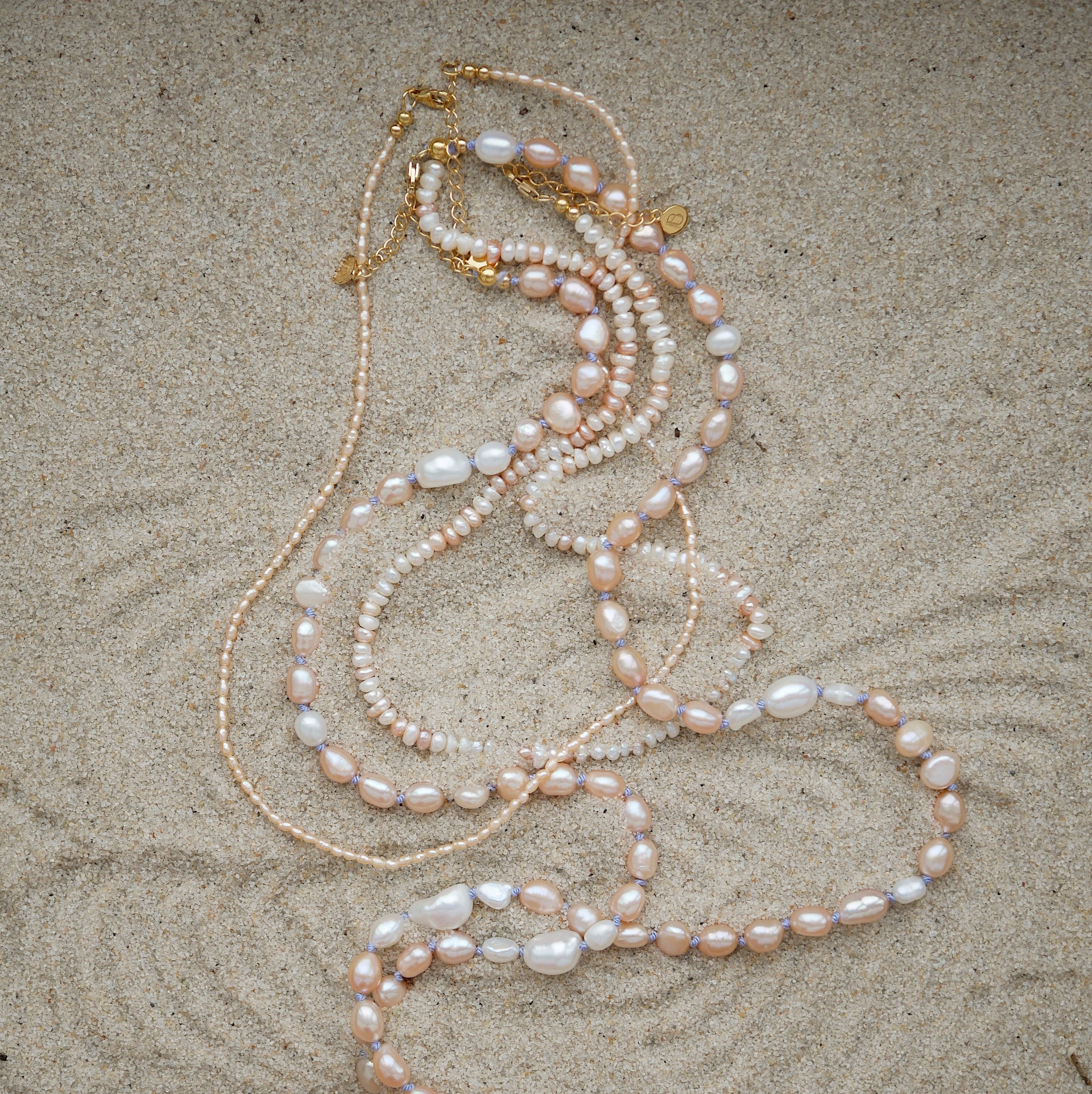 Pearl necklaces with pink, white, and cream pearls, accented with gold-tone chain and clasp, laid out on a sandy surface.