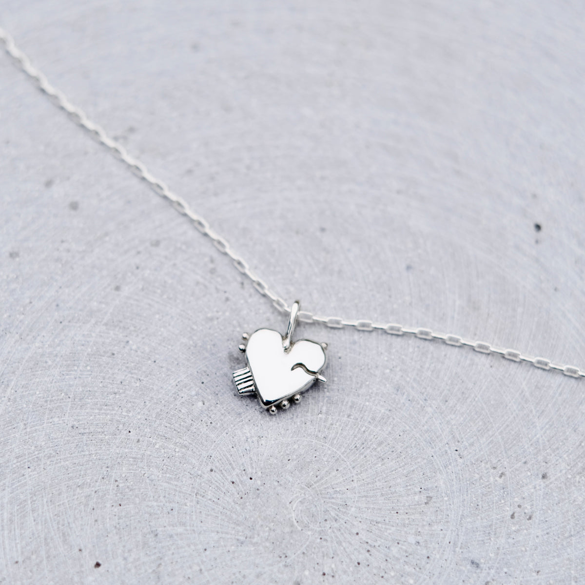 A delicate silver necklace with a small heart-shaped pendant resting on a textured light gray surface.
