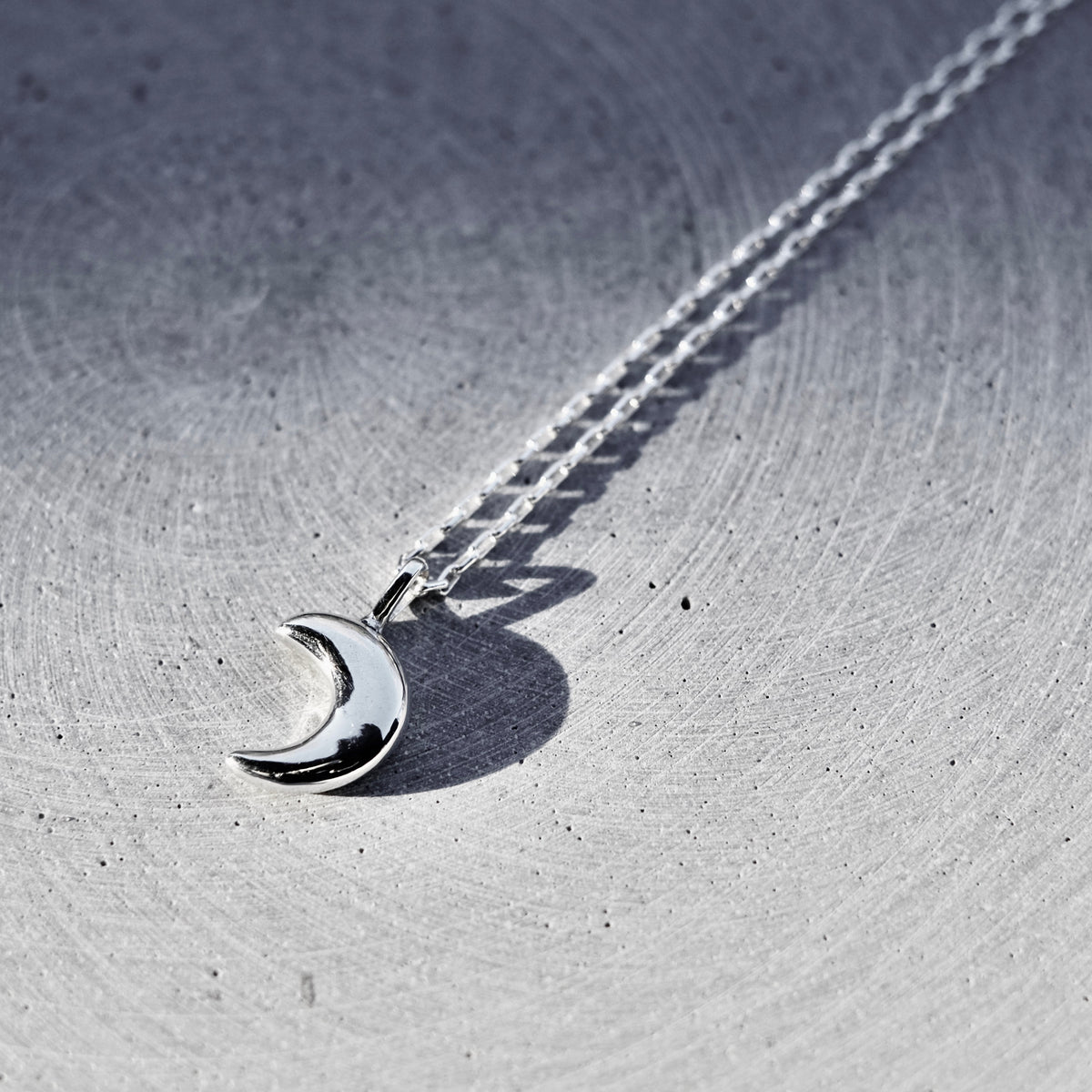 A silver crescent moon pendant necklace on a matching chain, displayed on a textured gray metallic surface.