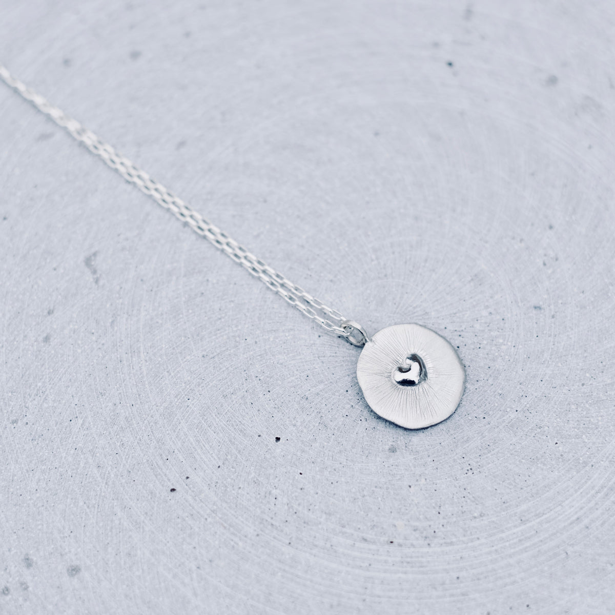 A delicate silver necklace with a small, flat circular disc pendant featuring a tiny heart engraving, resting on a textured light gray surface.