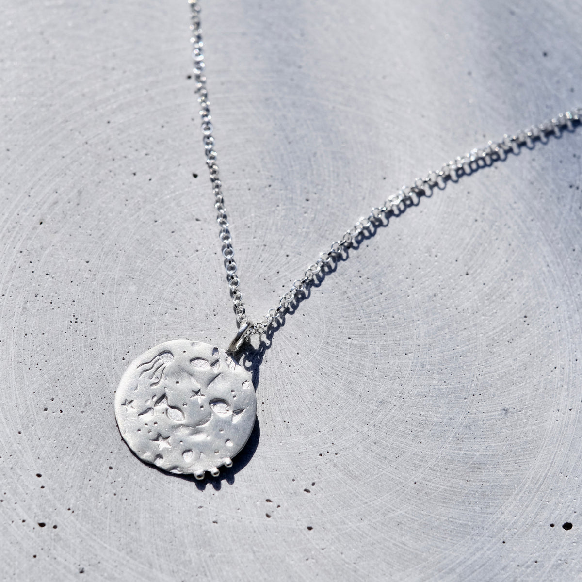 A silver necklace with a textured, circular moon-shaped pendant, displayed on a light gray concrete surface.