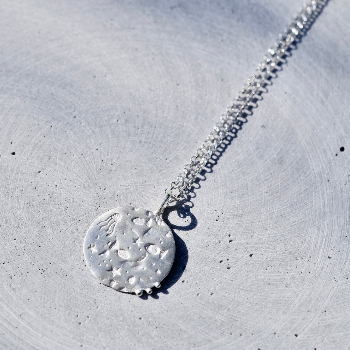 A silver pendant necklace with a textured, hammered circular disc pendant, resting on a light gray concrete surface.