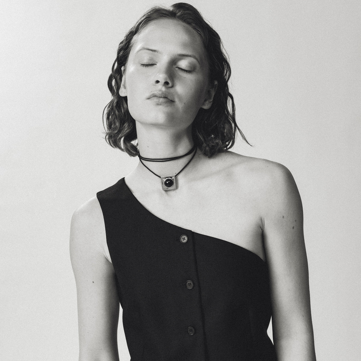 A black one-shoulder button-front vest worn by a model with her eyes closed, paired with a white skirt against a light gray studio background.