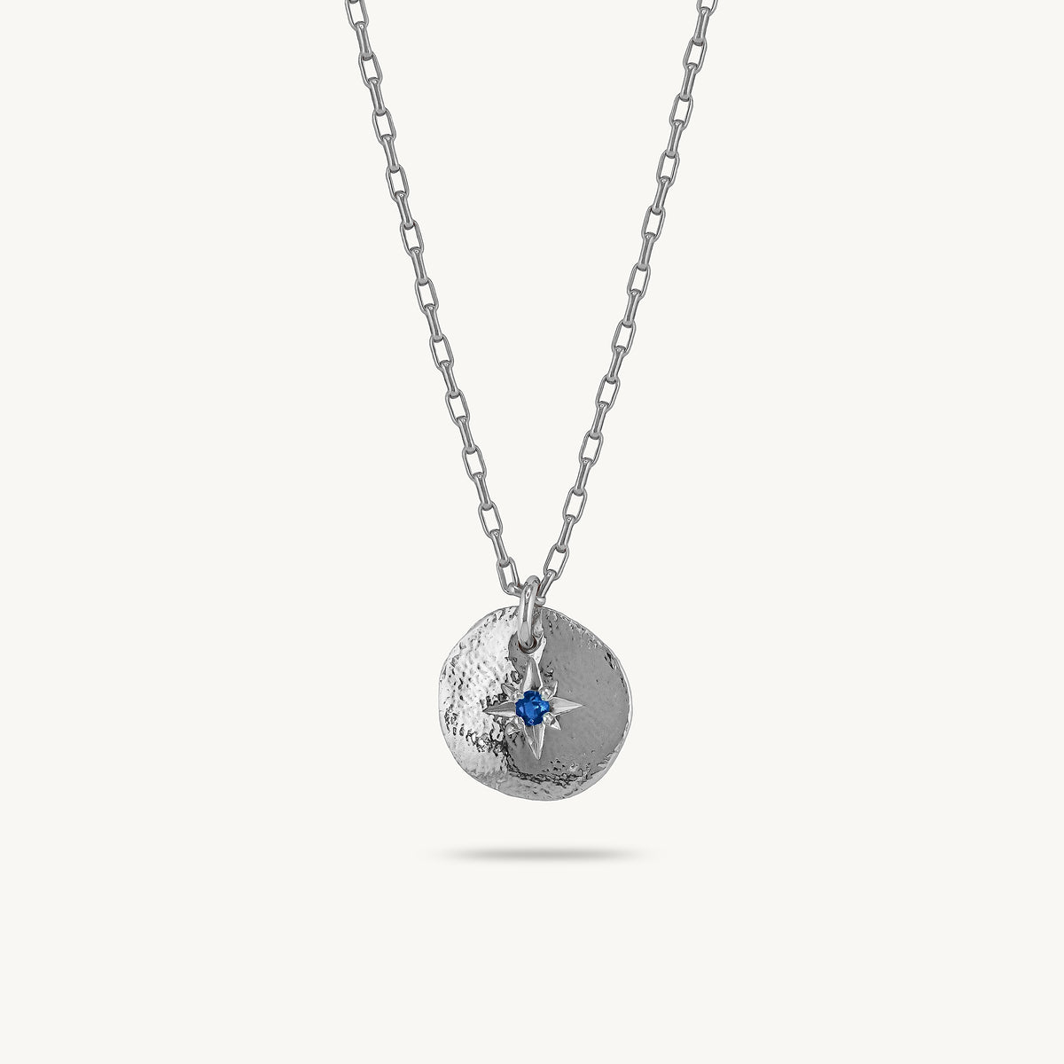 A silver chain necklace with a textured circular pendant featuring a small blue sapphire, shown against a light cream background.