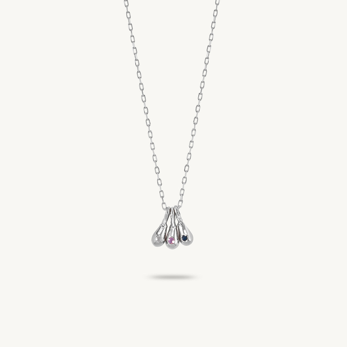 A delicate silver necklace with a pendant featuring three teardrop-shaped gold drops, one of which is set with a small iridescent stone, displayed against a light cream background.