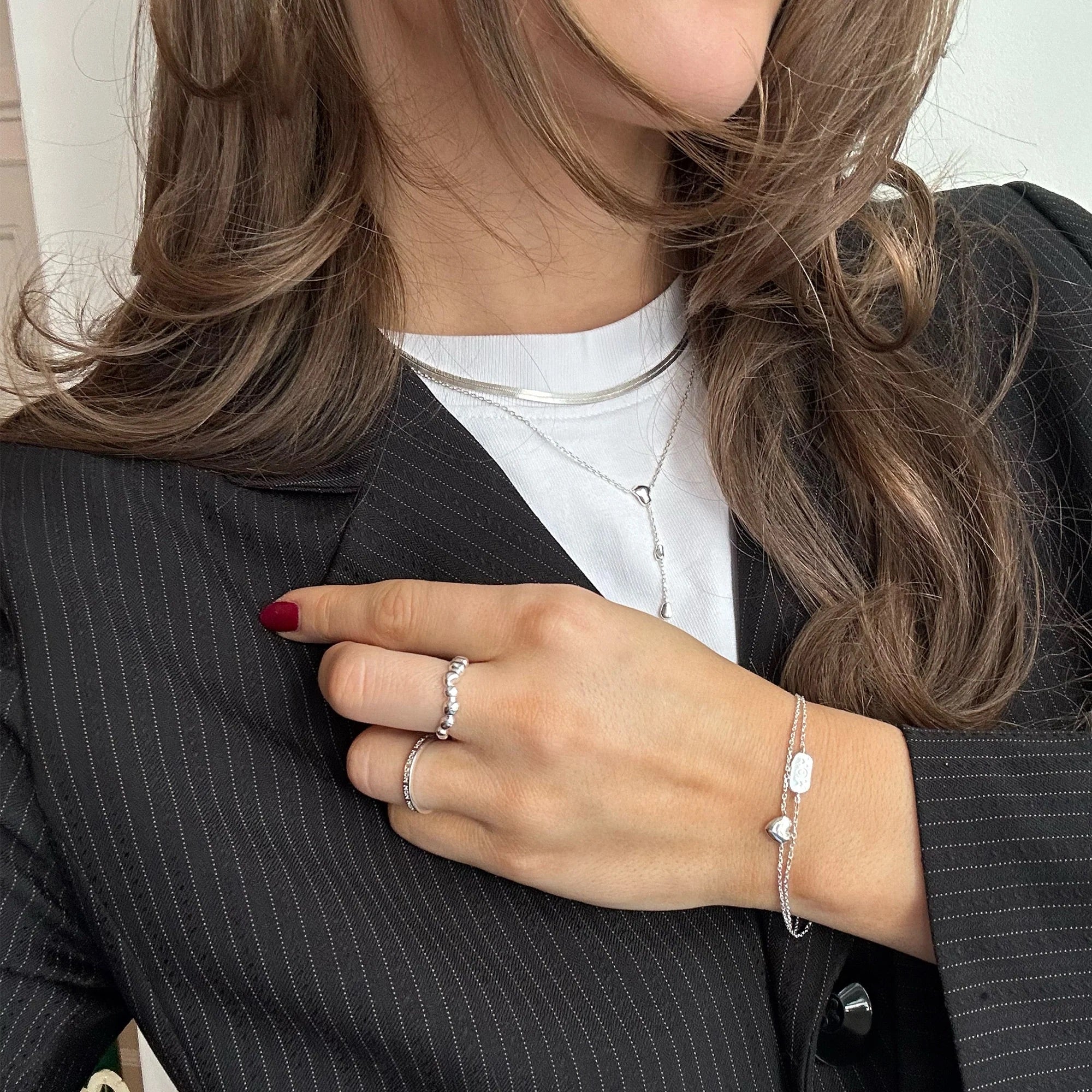 A close-up lifestyle shot of a woman wearing delicate silver jewelry, including a silver chain bracelet with white beads, silver rings, and a silver necklace, styled with a black pinstripe blazer and white t-shirt.
