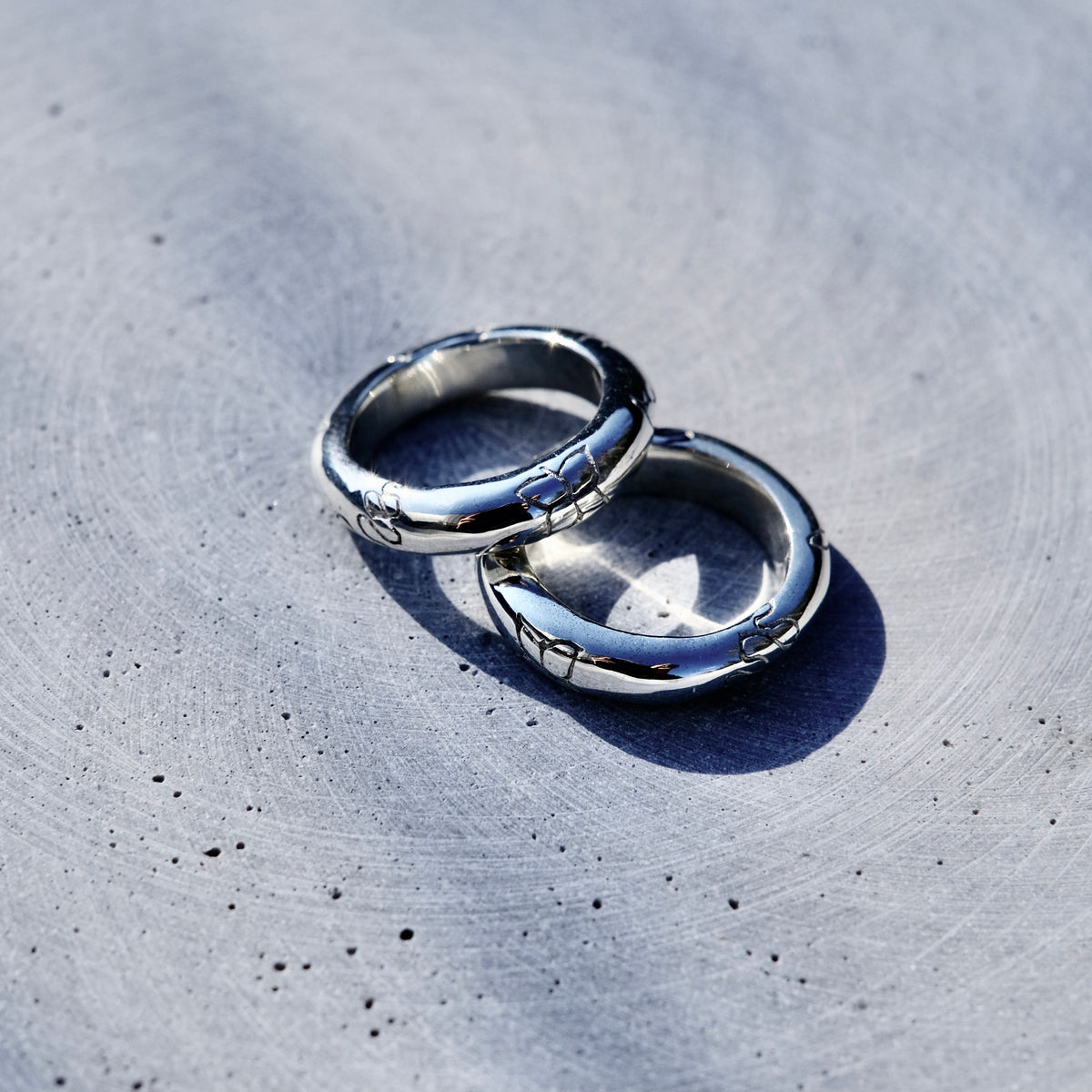 Two polished silver rings with engraved patterns, resting on a textured gray concrete surface under bright natural sunlight.