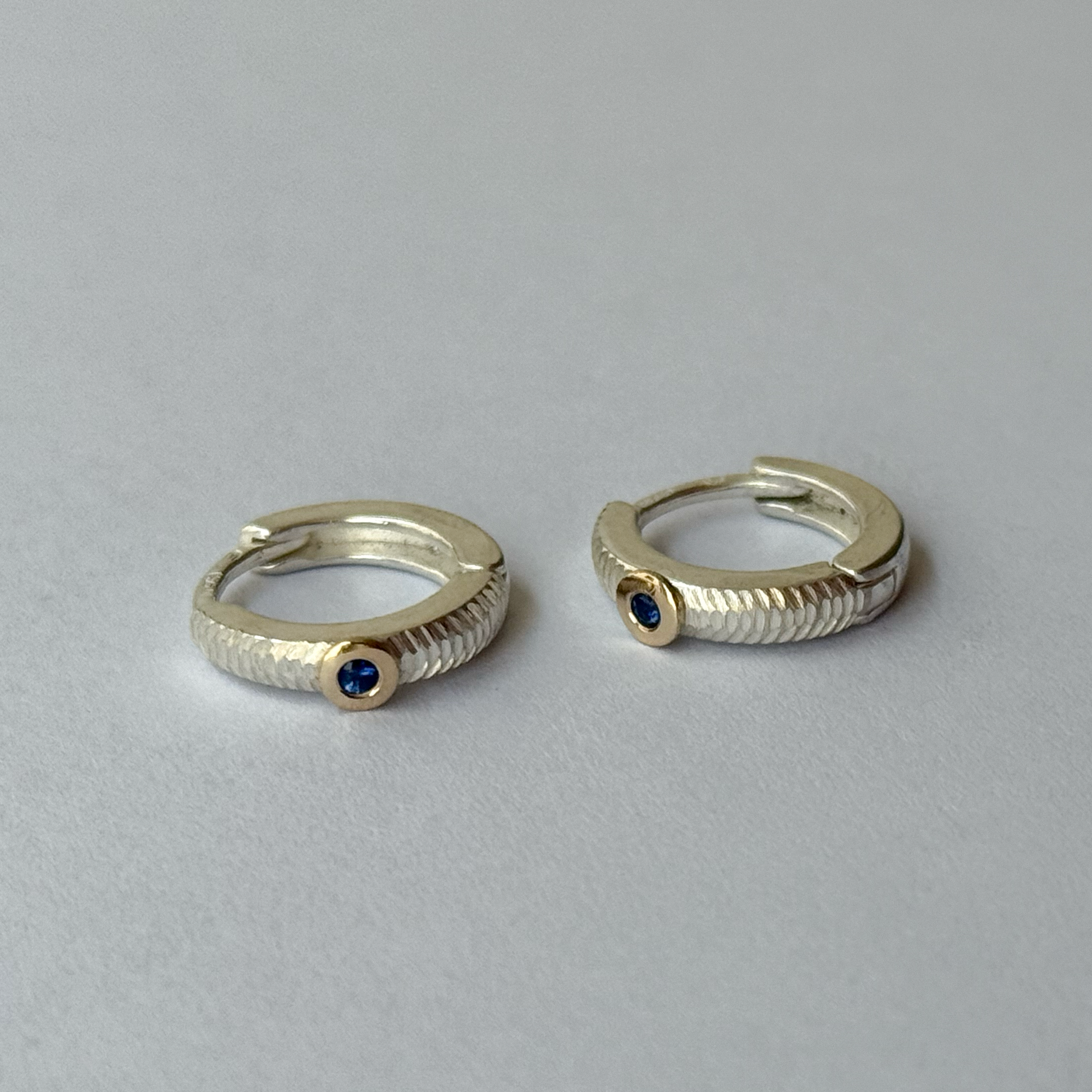 A pair of silver hoop earrings with blue sapphires and gold accents, placed on a white surface.