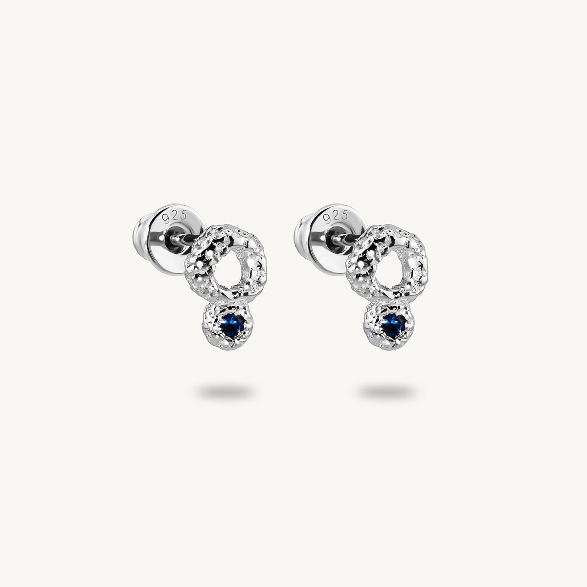 A pair of silver stud earrings with a textured open circular design and a small dark blue sapphire dangling below, set against a light cream background.
