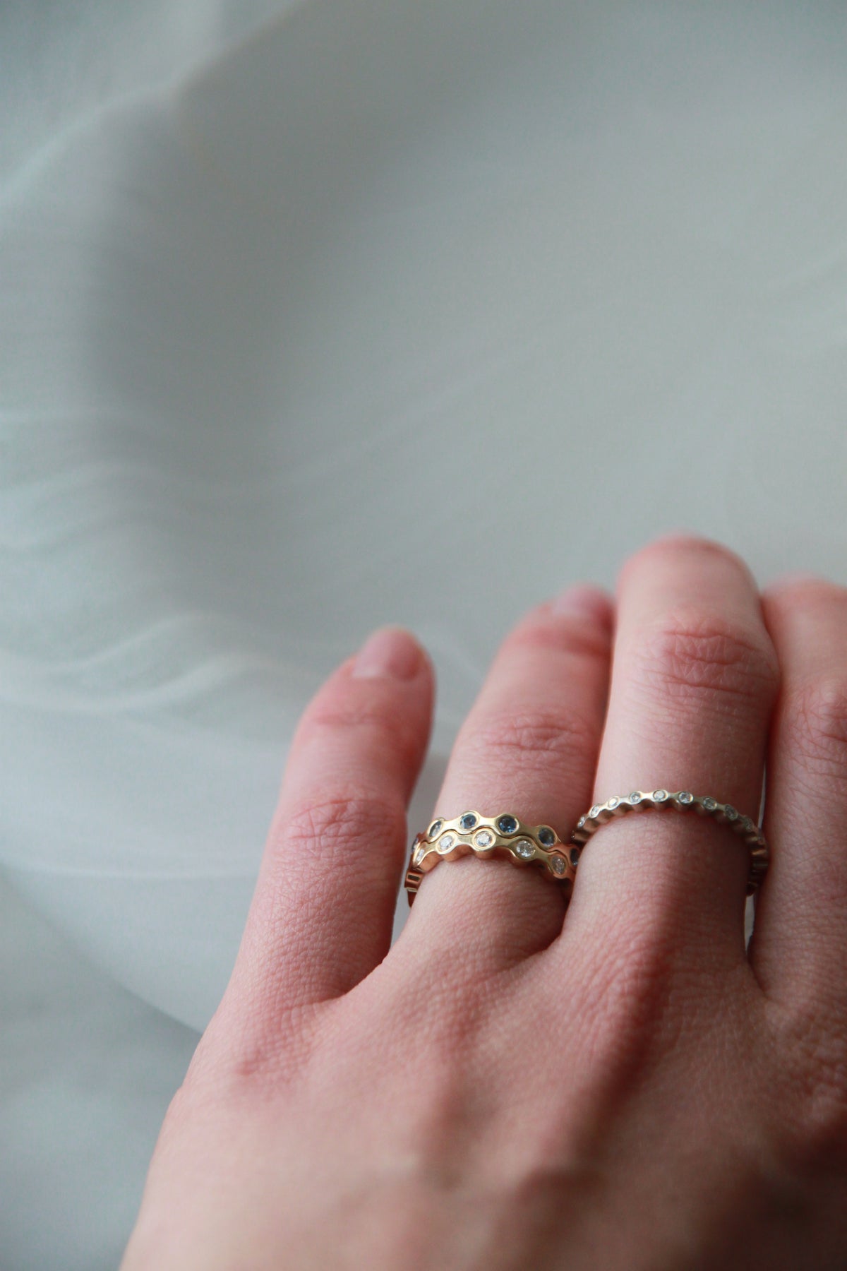 A close-up of a hand wearing two delicate rings: a gold band with circular cutout detailing and a thin silver band with small clear stones, set against a soft, light gray background.