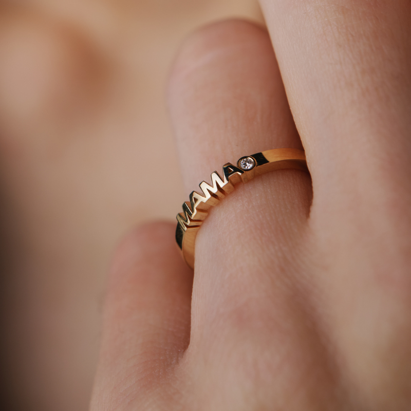 Gold ring with 'MAMA' word set with diamond on a finger against a blurred background