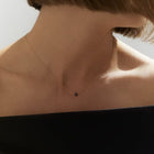 A close-up of a model's neck and upper chest wearing a delicate gold chain necklace with a small black faceted diamond, paired with a black off-the-shoulder top.