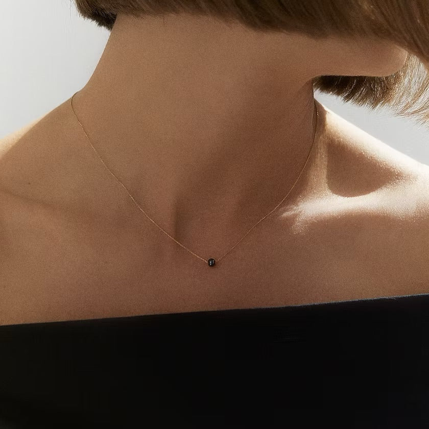 A close-up of a model's neck and upper chest wearing a delicate gold chain necklace with a small black faceted diamond, paired with a black off-the-shoulder top.