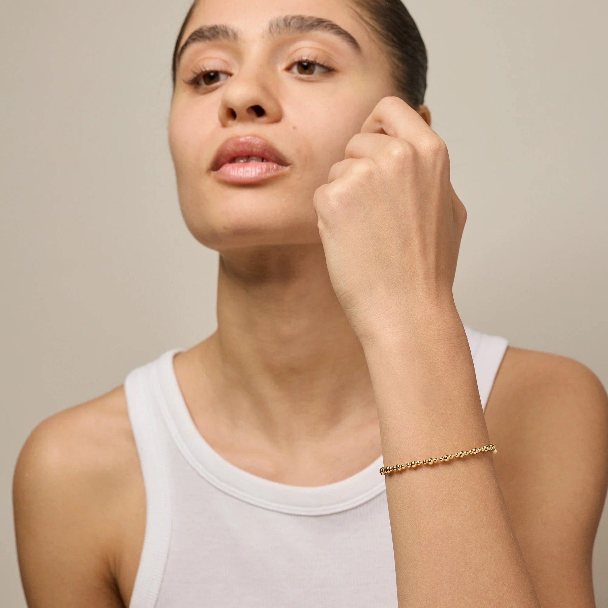 A close-up of a woman wearing a delicate gold chain bracelet on her wrist, paired with a white tank top against a neutral beige background.