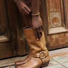 Brown leather boot with a decorative buckle worn by a person against a wooden door.