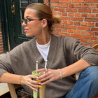 A woman wearing a taupe brown V-neck knit sweater over a white t-shirt and blue jeans, sitting outdoors in front of a red brick wall and holding a green drink.