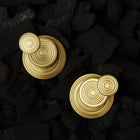 Two gold-toned circular layered earrings with concentric ring details, displayed on a bed of black charcoal.