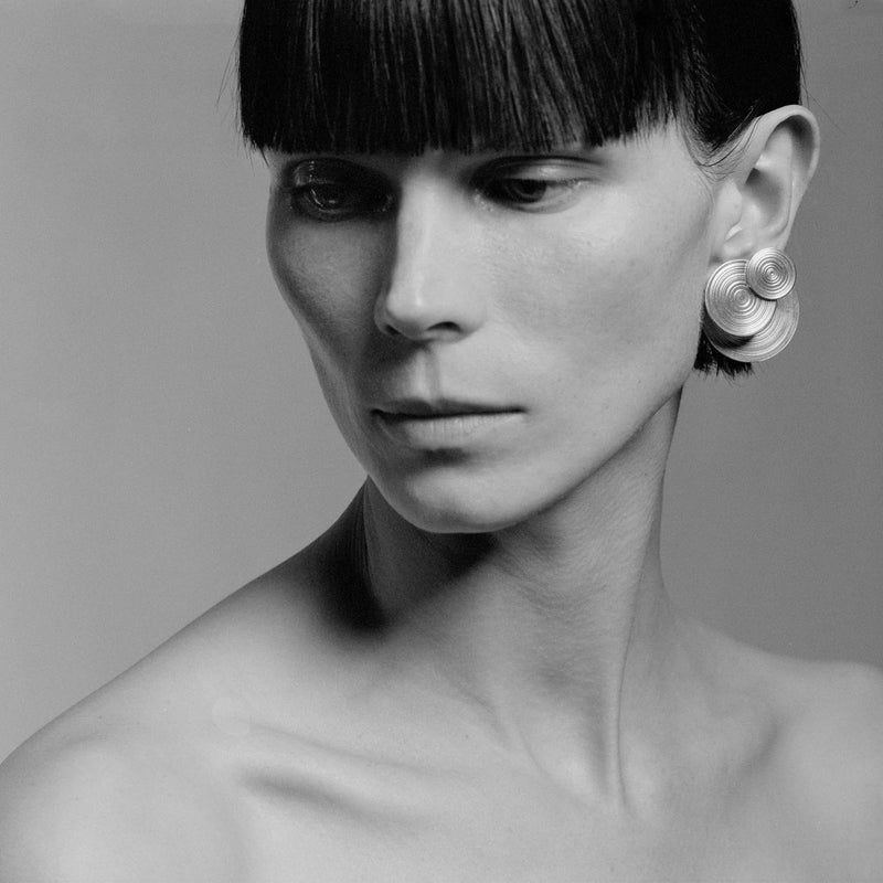 A close-up black and white portrait of a model with short dark hair and a large spiral-shaped silver earring, shown against a plain gray studio background.