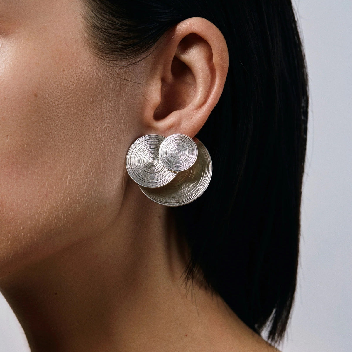 A close-up of a model's ear wearing a pair of large, silver spiral disc earrings against a light gray background.
