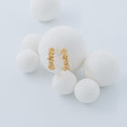 A pair of textured gold hoop earrings displayed on a large white foam ball, surrounded by smaller white foam balls against a light gray background.