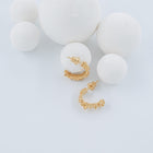 A pair of small gold huggie hoop earrings with a textured, floral-like pattern, displayed on a light blue background with white foam spheres.