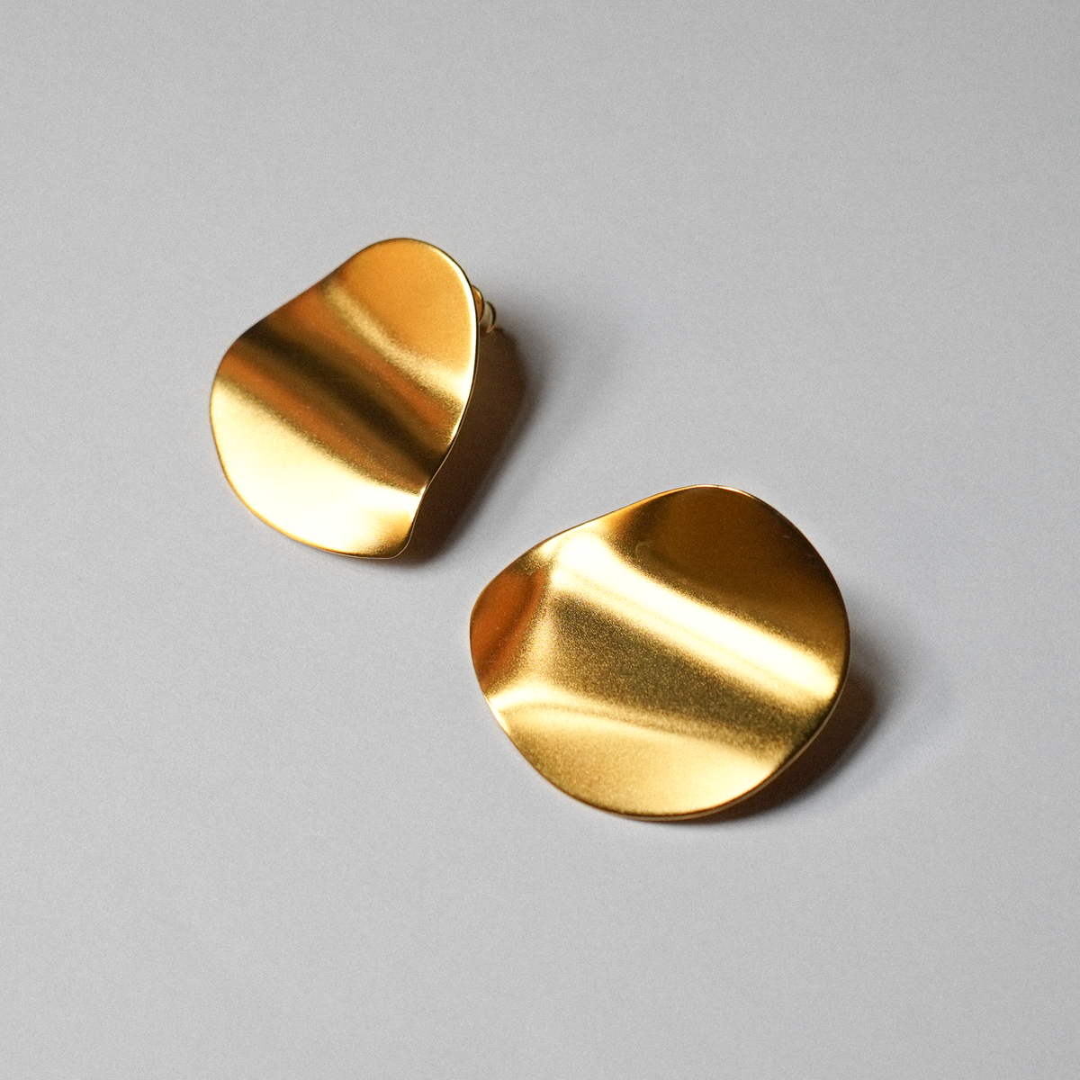 A pair of abstract, organic-shaped gold-plated silver earrings with a brushed metallic finish, displayed on a neutral light gray background.