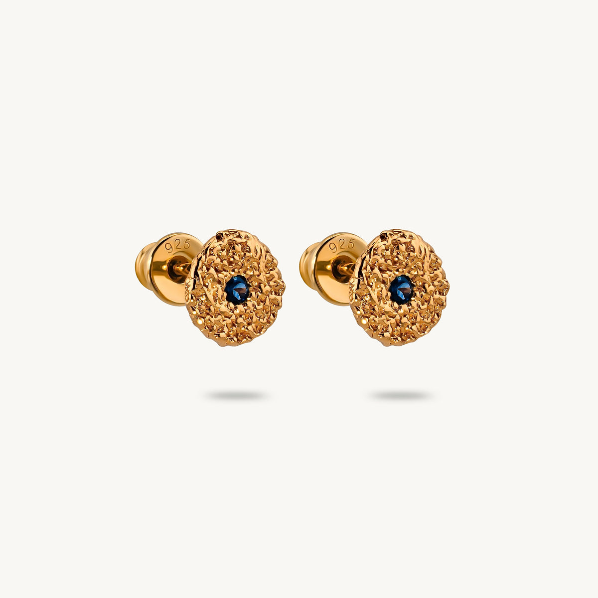 A pair of gold-plated silver stud earrings with a textured, flower-like circular face and a small round blue sapphires in the center, shown on a light off-white background.