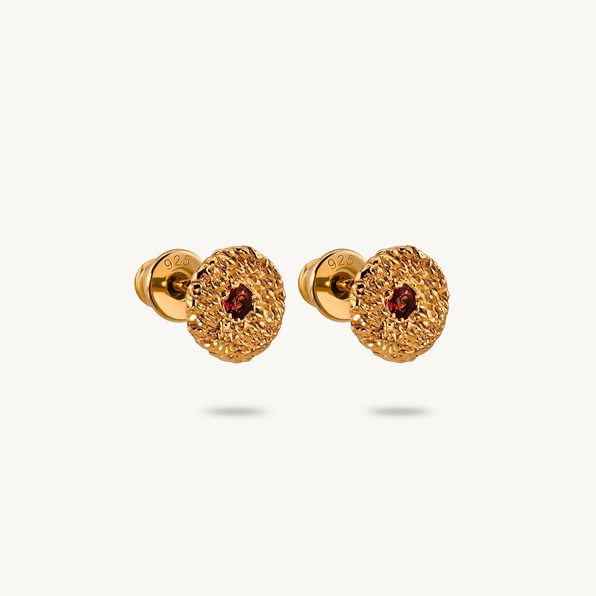 A pair of gold stud earrings with a textured, flower-like circular face and a small round red sapphires in the center, shown on a light off-white background.