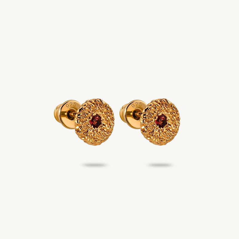 A pair of gold stud earrings with a textured, flower-like circular face and a small round red sapphires in the center, shown on a light off-white background.