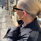 A close-up lifestyle shot of a woman outdoors wearing gold hoop earrings, black sunglasses, and a dark coat, with a cobblestone street in the background.