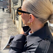 A close-up lifestyle shot of a woman outdoors wearing gold hoop earrings, black sunglasses, and a dark coat, with a cobblestone street in the background.