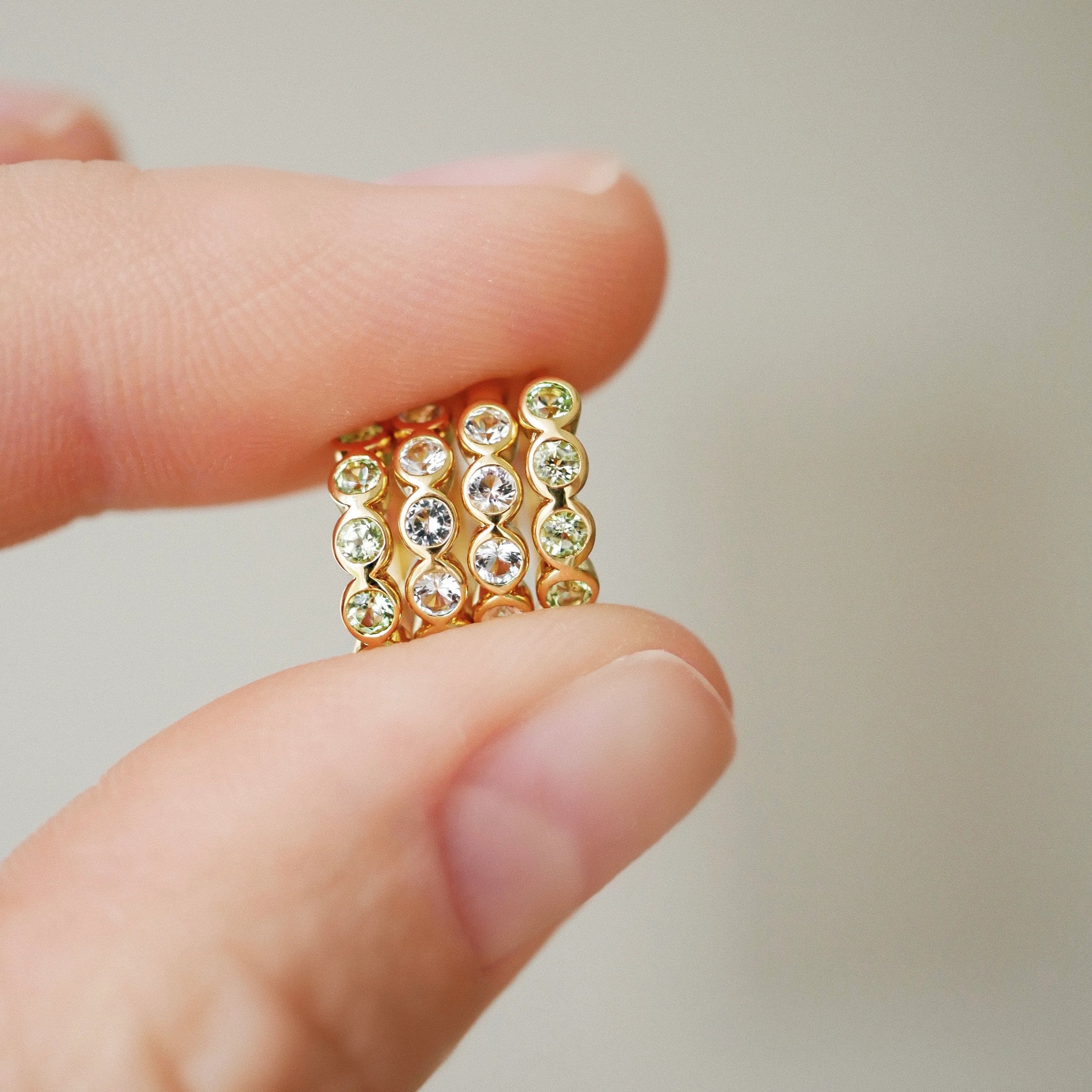 A close-up of a hand holding a stack of three gold eternity rings set with clear round-cut gemstones against a soft, neutral beige background.