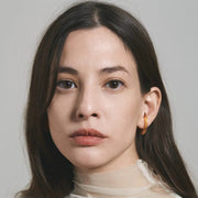 A close-up portrait of a woman with long dark brown hair, wearing a small gold hoop earring, against a plain light gray background.