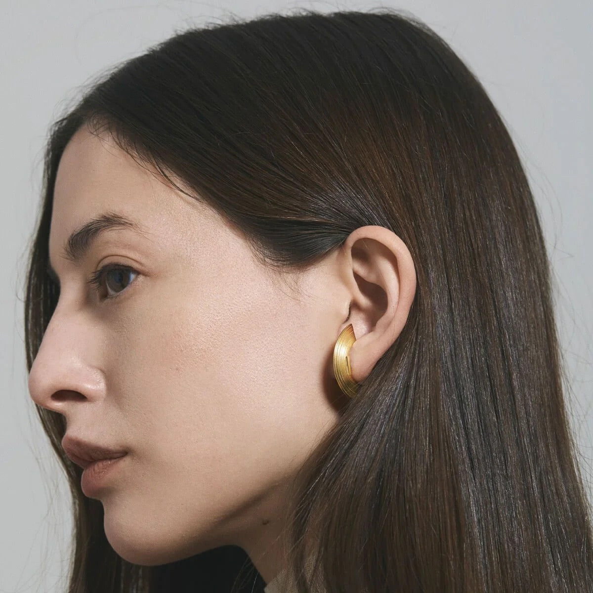 A close-up side profile of a woman with long dark brown hair wearing a single thick, polished gold hoop earring against a neutral light gray background.