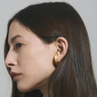 A close-up side profile of a woman with long dark brown hair wearing a single thick, polished gold hoop earring against a neutral light gray background.