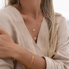 A close-up of a woman wearing layered gold necklaces with round and textured pendants, a delicate gold bracelet, and a beige linen wrap top.