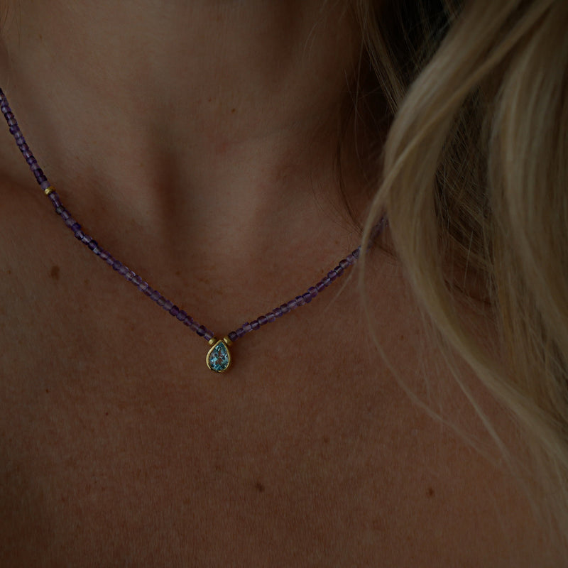 A close-up of a person's neck and chest wearing a delicate beaded necklace with a small gold teardrop pendant featuring a light blue stone.