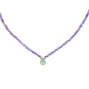 A delicate necklace featuring a strand of small purple amethyst beads and a central gold-tone pendant with a teardrop-shaped light blue gemstone, set against a plain white background.