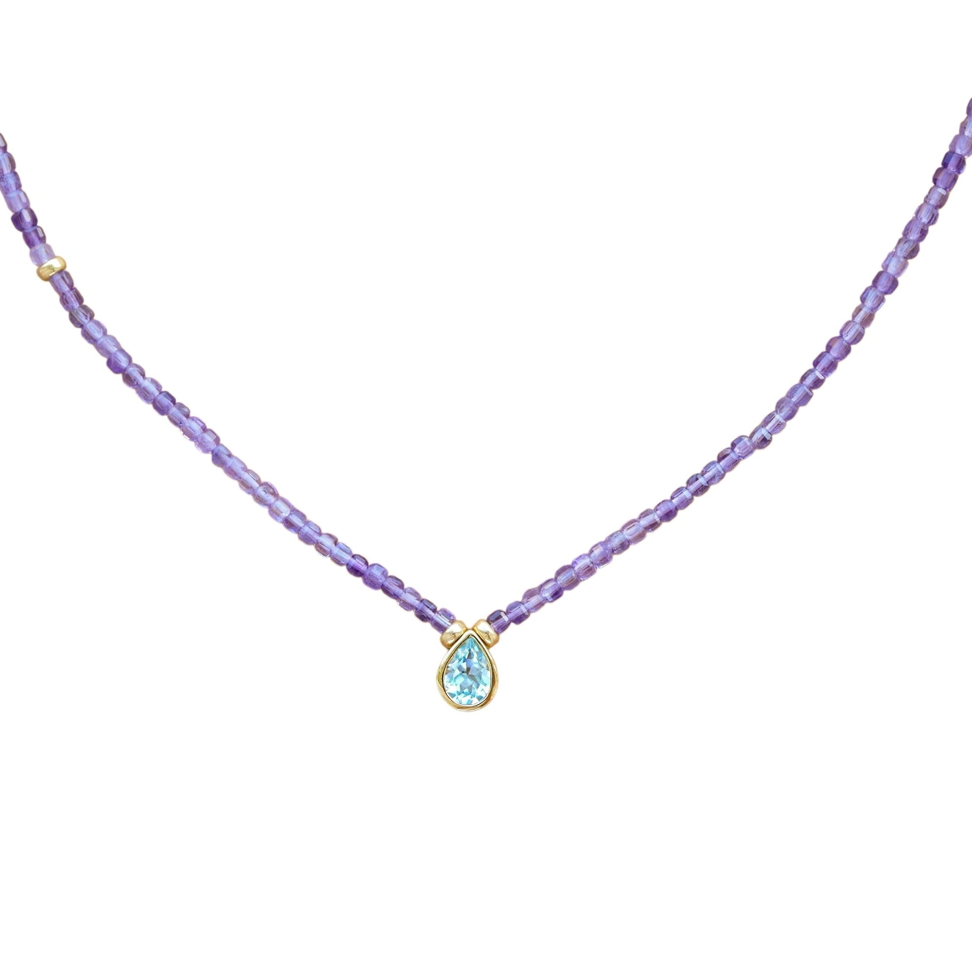 A delicate necklace featuring a strand of small purple amethyst beads and a central gold-tone pendant with a teardrop-shaped light blue gemstone, set against a plain white background.