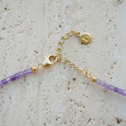 Close-up of a delicate gold-plated silver chain necklace extender with a lobster clasp, a small round branded charm, and purple faceted beads on a light textured stone surface.