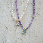 Two beaded necklaces displayed on a textured beige stone surface. One necklace features white pearl-like beads with a gold teardrop pendant containing a clear faceted stone.