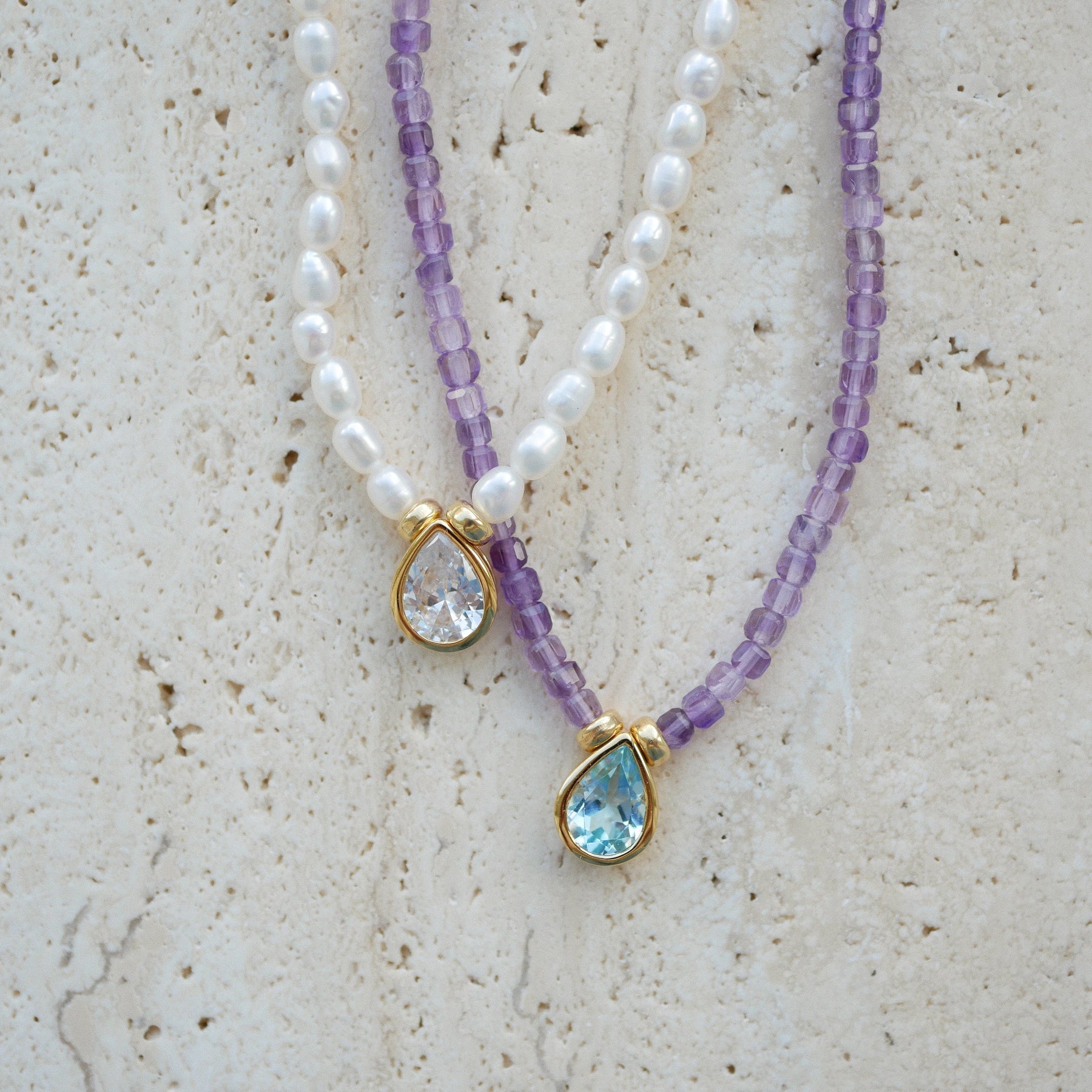 Two beaded necklaces displayed on a textured beige stone surface. One necklace features white pearl-like beads with a gold teardrop pendant containing a clear faceted stone.