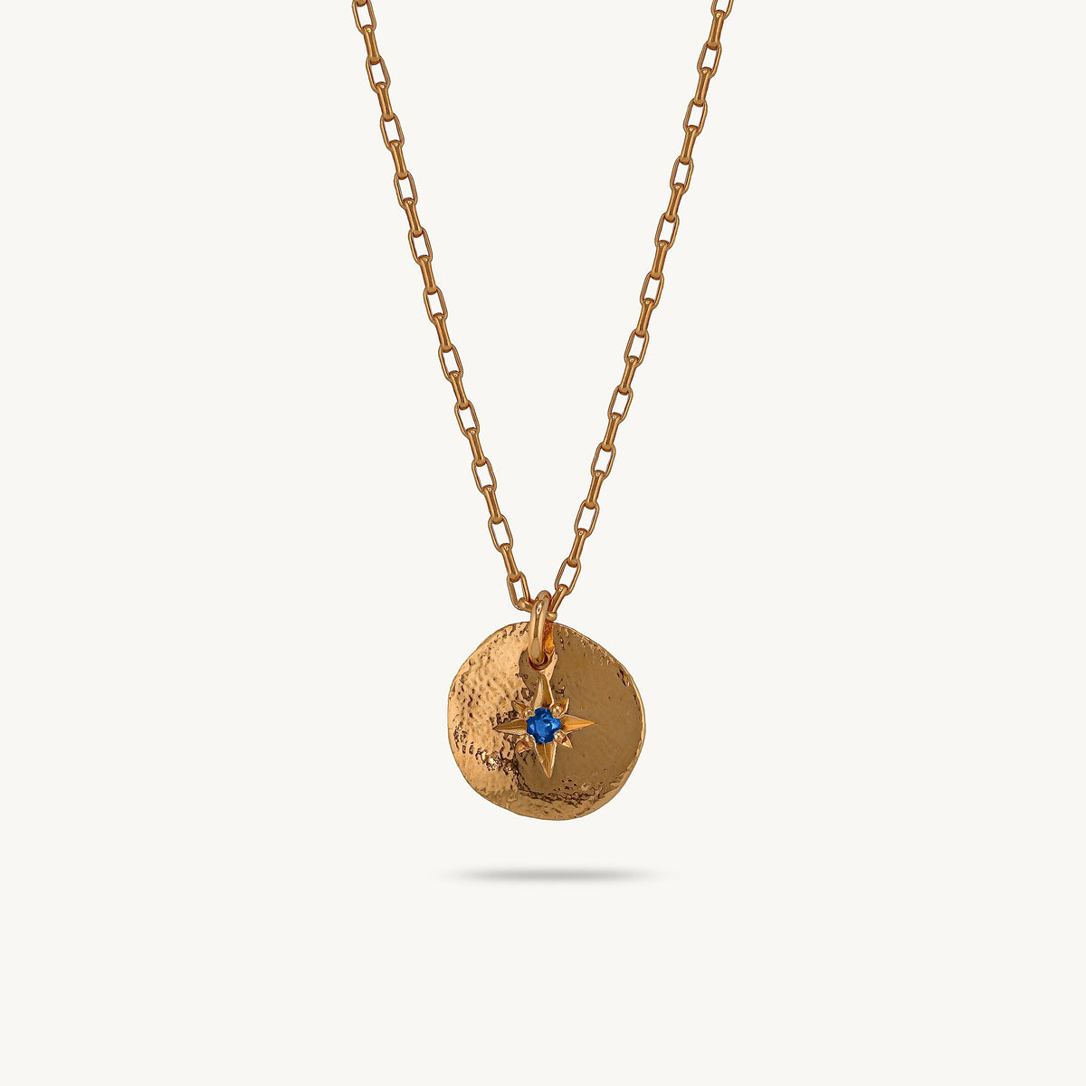 A gold-toned chain necklace with a textured circular disc pendant featuring a small blue sapphire gemstone and gold star detail, shown against a light off-white background.
